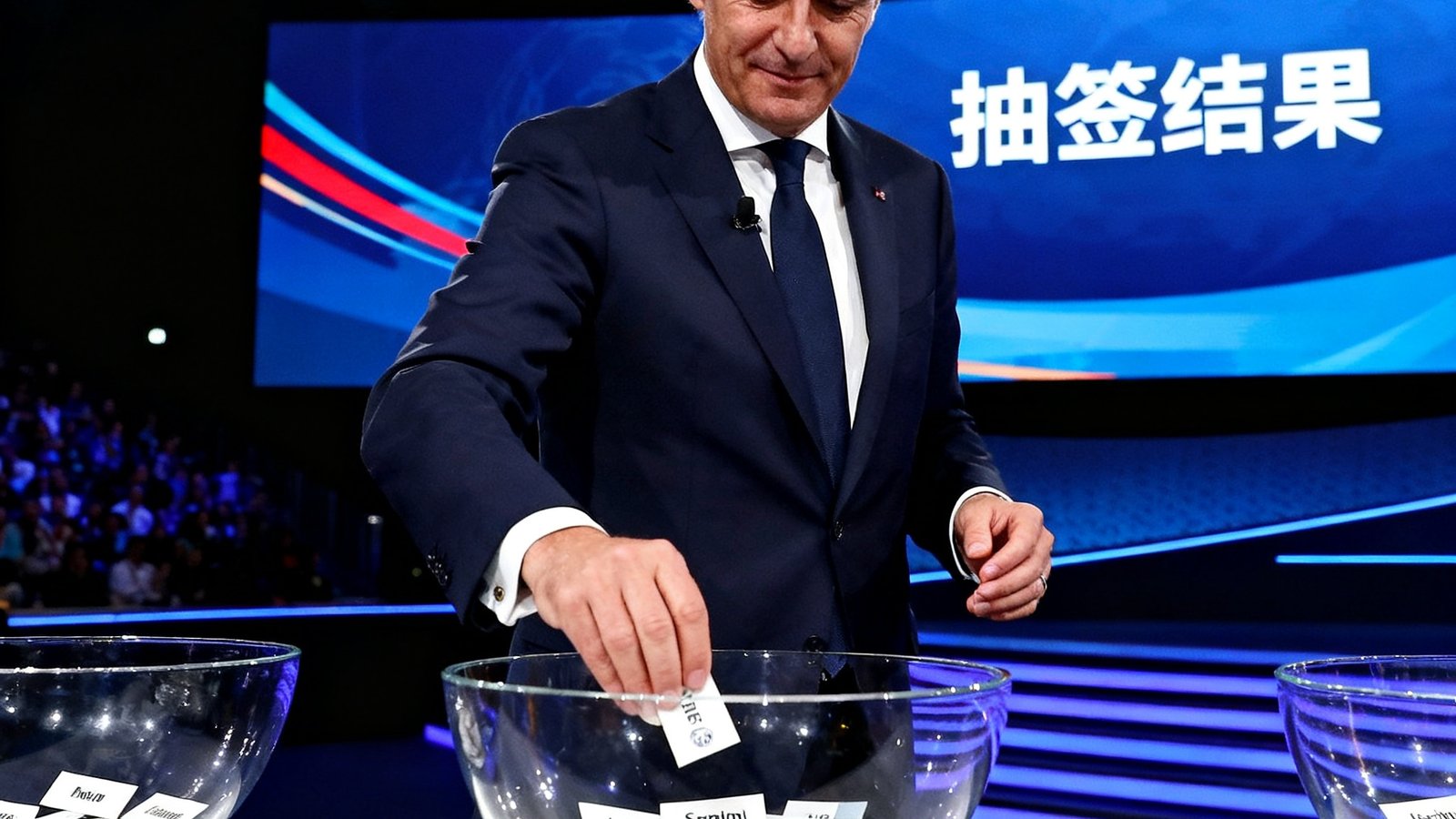 欧冠淘汰赛抽签仪式现场照片，欧足联官员站在舞台上从玻璃碗中抽出印有球队名称的名牌，大屏幕同步显示抽签结果，现场气氛紧张